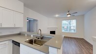 This two-floor residence offers a thoughtfully designed open-concept layout that seamlessly connects the kitchen and living areas, creating a functional and inviting atmosphere throughout. The kitchen is outfitted with crisp white cabinetry, a sleek quartz countertop with a peninsula that doubles as a breakfast bar, and a stainless steel undermount sink with a modern pull-down faucet — all contributing to a clean, contemporary aesthetic. The adjoining living area is warm and welcoming, anchored by a classic fireplace with a white surround that serves as a natural focal point. A ceiling fan overhead provides both comfort and a subtle architectural detail. Durable hardwood-style flooring runs throughout the main living space, adding warmth and visual continuity. A large window invites abundant natural light, offering pleasant outdoor views and brightening the interior considerably. A doorway visible from the kitchen suggests additional rooms and convenient flow between spaces. The combination of neutral tones, modern fixtures, and thoughtful layout makes this a practical yet stylish home suited for everyday living across its two levels.