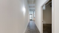 This one-bedroom, one-bathroom loft in Petersburg, Virginia offers a distinctive urban industrial aesthetic throughout its efficiently designed single-floor layout. Polished concrete floors run continuously across every room, lending a cohesive, modern character to the space. Soaring ceilings with exposed beams and structural elements amplify the sense of openness well beyond what the footprint might suggest.

The combined kitchen and living area serves as the heart of the unit. The kitchen is outfitted with rich, dark-stained cabinetry, granite-style countertops, black appliances, and a ceiling fan overhead — a practical yet stylish arrangement. The adjacent living space benefits from large, industrial-style windows that flood the area with natural light and provide views of the surrounding neighborhood.

A hallway connects the main living area to the private quarters, featuring wall sconce lighting and clean white walls. The bedroom is a well-proportioned retreat with soaring ceilings, a high transom-style window for added natural light, and two closets with natural wood doors. The bathroom is clean and functional, equipped with a full tub/shower combo. A dedicated laundry area adds everyday convenience. The overall ambiance is that of a refined urban loft with a minimalist, industrial character.