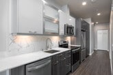 This contemporary single-floor urban residence showcases a thoughtfully designed layout with modern finishes throughout. The galley-style kitchen is efficiently organized, featuring floor-to-ceiling dark wood cabinetry, stainless steel appliances including a full-size refrigerator, dishwasher, and microwave, complemented by a white quartz countertop with a marble-effect backsplash. A distinctive exposed concrete structural column adds an industrial character to the kitchen and living area, contrasting beautifully with the clean, neutral palette of the surrounding walls. Hardwood-style flooring in warm and cool tones flows consistently throughout the space, lending a cohesive and polished aesthetic. The living and bedroom areas are bathed in natural light through expansive floor-to-ceiling windows that frame sweeping city skyline views, bringing the urban landscape directly into the home. The bathroom is finished to a high standard, featuring a backlit LED vanity mirror, a white undermount sink with chrome fixtures, and a glass-enclosed shower adorned with elegant marble-style tile. Recessed lighting throughout ensures the space is well-illuminated, while the open and clean design creates a bright, airy atmosphere ideal for modern city living.