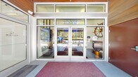 Located at 2723 California Ave SW in Seattle's West Seattle neighborhood, Admiral Station's ground-floor lobby presents a well-appointed communal space designed to serve residents with both functionality and visual appeal. The generously sized single-floor layout accommodates several distinct functional areas that flow seamlessly into one another.

The main lobby entrance features large glass double doors that flood the interior with natural light, opening into a polished concrete-floored reception area. A signature architectural detail is the raised tray ceiling, illuminated with vibrant blue ambient lighting and anchored by stylish industrial-style pendant fixtures, creating a striking focal point overhead. White walls adorned with nautical-themed artwork and motivational decor reinforce the building's Admiral Station identity.

Resident mailboxes—both traditional and smart parcel lockers—line the walls, providing convenient mail retrieval. A staffed management office with glass partitions sits adjacent to the lobby, accompanied by a small seating area. A stainless steel elevator offers access to upper floors. A dedicated fitness room with glass doors and teal accent walls houses exercise equipment. A utility corridor with office storage and service cart access completes the functional back-of-house areas.