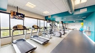 This fitness center is a thoughtfully designed, single-floor facility that offers a comprehensive range of workout equipment within a bright, modern environment. The space is defined by its bold teal accent walls and ceilings, which contrast sharply with the neutral white tile ceiling and light-toned speckled flooring, creating an energetic yet clean aesthetic throughout.

Large floor-to-ceiling windows line one full wall of the facility, flooding the interior with natural light and offering expansive views of a treetop canopy and open waterway beyond — a distinctive backdrop that elevates the workout experience. Multiple flat-screen televisions are mounted throughout the space, providing entertainment for cardio sessions.

The cardio zone features a well-organized row of treadmills, elliptical machines, stationary bikes, and rowing machines, all positioned to take advantage of the outdoor views. A dedicated strength training area includes a cable/functional trainer machine, a full rack of kettlebells in graduated sizes, a BOSU balance trainer, and a weight rack, offering versatile options for resistance and functional training. Exposed industrial ductwork along the ceiling adds an intentional design element, complementing the gym's modern character.

The overall layout is open and spacious, promoting ease of movement between zones, while the abundant natural light and scenic views make this fitness center an inviting and motivating space for daily exercise.
