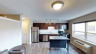 This single-floor space showcases a thoughtfully designed open-concept layout that seamlessly connects the kitchen and living areas. The kitchen serves as the focal point of the space, featuring rich dark-stained cabinetry that extends to the upper walls, providing ample storage. Granite countertops add a refined touch, complemented by a full suite of stainless steel appliances including a refrigerator, dishwasher, gas range, and built-in microwave. A movable kitchen island with a matching granite surface and dark base offers flexible additional workspace and casual dining potential. Track lighting above the kitchen and a flush-mount ceiling fixture ensure the space is well-lit throughout. Light-toned hardwood-style flooring runs throughout, lending warmth and continuity to the open floor plan. A window allows natural light to filter into the space, enhancing the bright and airy atmosphere. A hallway visible in the background leads to additional areas of the unit, including what appears to be a closet or utility space with a door. The overall design balances functionality with modern aesthetics, making it a practical and visually appealing living environment.