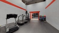 This fitness facility is organized across a single floor and divided into distinct training zones connected by striking red-framed doorways that serve as both functional passages and bold design statements. A consistent visual identity is established throughout the space via red horizontal accent stripes running along the upper walls, contrasting against a neutral grey and white color scheme.

The cardio zone is a long, corridor-style room lined with treadmills, air bikes, and rowing machines arranged along one wall. Diamond-plate rubber flooring provides a durable, slip-resistant surface, while a wall-mounted flat-screen television offers entertainment during workouts. Overhead panel lighting ensures the area is well illuminated.

Adjacent to the cardio zone is a strength and functional training area featuring high ceilings and an open layout. This space is equipped with a squat rack, a tiered dumbbell rack, a collection of kettlebells in varying weights, and medicine balls organized on a dedicated rack. A large mirror panel enhances the sense of space and supports form monitoring. Wall-mounted speakers support an energetic workout atmosphere.

The flooring transitions from rubber tile in the training areas to light hardwood in connecting zones, reinforcing the separation between functional spaces while maintaining a cohesive overall aesthetic.