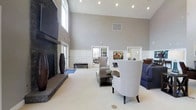 This thoughtfully designed single-floor space exudes a refined yet inviting ambiance, highlighted by dramatic soaring ceilings that amplify the sense of openness and grandeur. The main lounge area serves as the centerpiece, anchored by a striking floor-to-ceiling stone fireplace paired with a large wall-mounted television, making it both a cozy and functional gathering point. Recessed lighting throughout the ceiling provides warm, even illumination, complementing the natural light that filters in through multiple windows. The space features classic white wainscoting along the lower walls, adding a touch of timeless architectural detail against the soft gray upper walls. Comfortable seating — including neutral-toned armchairs, a navy sofa, and a dark wood coffee table — is arranged to encourage conversation and relaxation. French-style glass-paned doors lead to adjacent rooms, offering a sense of connectivity and flow throughout the space. Plush wall-to-wall carpeting in a neutral tone ties the entire area together, contributing to its warm and welcoming character. Colorful artwork adds a vibrant finishing touch, making this a polished and practical space well-suited for lounging, socializing, or unwinding.