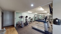 Located at 260 Rim View Dr in Colorado Springs, Colorado, this fitness center is a well-appointed, single-floor facility designed to support a comprehensive workout experience. The space features a clean, modern interior with a combination of carpet tile and hardwood-style flooring, recessed and fluorescent lighting, and a neutral color palette accented by a signature mint-green feature wall.

The cardio zone is equipped with multiple treadmills, elliptical trainers, and stationary bikes, providing ample options for aerobic exercise. A wall-mounted television is positioned to keep users entertained during cardio sessions. The strength training area includes a multi-station cable machine, a weight bench, and a well-stocked dumbbell rack with kettlebells, catering to a range of resistance training needs. Floor-to-ceiling mirrors along one wall enhance the sense of space and allow users to monitor their form.

Motivational typography — "Don't Wish For It, Work For It" — is prominently displayed on the feature wall, reinforcing an energetic atmosphere. A rock climber silhouette mural adds an adventurous, Colorado-inspired aesthetic touch. A water fountain station is conveniently located near the exit. The facility is efficiently organized to maximize usable floor space while maintaining clear movement pathways throughout.