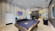 This well-appointed single-floor amenity space blends modern design with functional comfort across several distinct zones. The centerpiece of the space is a generously sized billiards area, anchored by a full pool table with a rich wood frame and navy felt surface. A bold, nature-inspired wallpaper featuring an abstract tree motif serves as a striking backdrop, complemented by a wall-mounted flat-screen TV and built-in bench seating with colorful accent cushions. Recessed lighting illuminates the area throughout.

An arched opening leads into an adjacent lounge area outfitted with comfortable seating, additional televisions, and sophisticated upholstered wall paneling, creating an inviting space for socializing and relaxation. A pendant chandelier adds an elegant focal point to this zone.

The space also includes a bright, open reception or common area with large-format tile flooring in a herringbone pattern, exposed brick accents, and curated wall art. Natural light filters in through multiple windows, enhancing the airy atmosphere.

A quiet nook near the windows offers a rustic wood worktable with industrial-style chairs — ideal for focused tasks. Throughout the space, the design balances contemporary finishes with warm textures, creating an environment suited to both leisure and productivity.