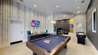 This well-appointed single-floor amenity space blends modern design with functional comfort across several distinct zones. The centerpiece of the space is a generously sized billiards area, anchored by a full pool table with a rich wood frame and navy felt surface. A bold, nature-inspired wallpaper featuring an abstract tree motif serves as a striking backdrop, complemented by a wall-mounted flat-screen TV and built-in bench seating with colorful accent cushions. Recessed lighting illuminates the area throughout.

An arched opening leads into an adjacent lounge area outfitted with comfortable seating, additional televisions, and sophisticated upholstered wall paneling, creating an inviting space for socializing and relaxation. A pendant chandelier adds an elegant focal point to this zone.

The space also includes a bright, open reception or common area with large-format tile flooring in a herringbone pattern, exposed brick accents, and curated wall art. Natural light filters in through multiple windows, enhancing the airy atmosphere.

A quiet nook near the windows offers a rustic wood worktable with industrial-style chairs — ideal for focused tasks. Throughout the space, the design balances contemporary finishes with warm textures, creating an environment suited to both leisure and productivity.