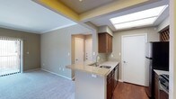 This single-floor space showcases a thoughtfully designed open-concept layout that integrates the kitchen and living area into one cohesive environment. The kitchen is a standout feature, equipped with rich dark wood cabinetry that provides ample storage, complemented by light granite countertops that add a touch of elegance. A peninsula with a breakfast bar extends outward, offering additional workspace and a casual dining option. The undermount sink is positioned along the peninsula, making it both functional and visually appealing.

A notable architectural highlight is the skylight positioned above the kitchen area, drawing in natural light and creating an airy, open atmosphere. The kitchen transitions into hardwood flooring, while the adjacent living area features plush carpeting, clearly defining each zone while maintaining an open feel. Neutral-toned walls throughout the space provide a versatile backdrop suitable for a variety of interior styles.

The layout also reveals a hallway leading to additional rooms, with doors visible in the background suggesting private spaces beyond. The overall design balances practicality with comfort, making it a functional and inviting living environment.