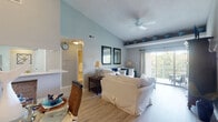Located in Coconut Creek, Florida, this single-story residence offers a well-designed open floor plan that seamlessly connects the living, dining, and kitchen areas. The living room is a standout feature, boasting soaring vaulted ceilings that amplify the sense of space, soft blue-gray walls, and light wood-style flooring. A ceiling fan overhead and a sliding glass door leading to a private balcony allow for natural light and outdoor views, enhancing the airy ambiance.

The dining area sits adjacent to the living room and is anchored by a pendant light fixture, while a pass-through window connects it to the kitchen. The kitchen is well-appointed with white cabinetry, quartz-style countertops, a tile mosaic backsplash, and stainless steel appliances including a refrigerator, range, and microwave.

Two bedrooms provide comfortable retreats, each featuring plush carpeting, natural light from large windows, and ample closet space—one with mirrored sliding doors and another with a walk-in closet. Two bathrooms serve the home, each with a shower/tub combination and vanity. The textured ceilings throughout add a classic Florida residential character to the overall space.