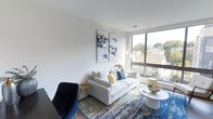 This single-floor space showcases a thoughtfully designed living area that balances modern aesthetics with functional comfort. The room is anchored by a large floor-to-ceiling window that floods the interior with abundant natural light while offering open views of the surrounding neighborhood, including mature tree canopies and neighboring rooftops. The crisp white walls enhance the sense of brightness and airiness throughout the space.

The living area is furnished with a contemporary white sofa accented by blue and yellow throw pillows, complemented by a sleek round white coffee table. A bold navy accent chair and a small gold side table add contrast and visual interest. Underfoot, a richly patterned area rug in deep blue and gold tones grounds the seating arrangement and introduces warmth to the hardwood flooring beneath.

Wall art featuring abstract blue and yellow compositions ties the color scheme together cohesively. The overall ambiance is clean, modern, and inviting, with a carefully curated palette that creates a polished yet welcoming atmosphere. The elevated position of the unit, suggested by the window views, adds an appealing sense of openness to the space.