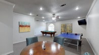 This single-floor game room is purpose-built for recreation and entertainment, offering a well-organized layout that accommodates multiple activities simultaneously. The space is anchored by a full-size pool table with a classic green felt surface, positioned beneath an arched window that allows natural light to filter in and brighten the room. Adjacent to it sits a regulation-size blue ping-pong table, providing a second focal point for competitive play.

At the center of the room, a round hammered-copper accent table is surrounded by houndstooth-patterned chairs, creating a comfortable casual seating area ideal for socializing between games. The neutral gray carpeting runs throughout, lending a cohesive and comfortable feel underfoot.

The walls are painted in crisp white and adorned with bold, colorful abstract artwork that adds visual interest and a lively energy to the space. Recessed lighting is paired with a ceiling fan at the center, ensuring the room remains well-lit and comfortably ventilated. Crown molding along the upper walls adds a refined architectural detail. A small tiled anteroom is visible at the entry, completing the functional and thoughtfully designed layout of this dedicated entertainment space.