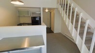 This two-story space offers a functional and straightforward layout designed for efficient living. The lower level features an open-concept design that integrates the kitchen and living area seamlessly. The kitchen is equipped with black appliances, including a refrigerator, and features a sink with upper cabinetry finished with frosted glass doors. A raised counter divides the kitchen from the adjacent living space, which also showcases a stainless steel surface, adding an industrial touch to the interior. A baseboard heating unit runs along the wall, providing climate control throughout the lower level. The flooring on the main level consists of neutral-toned carpet, contributing to a clean and understated aesthetic. White walls throughout maintain a bright and open atmosphere. A white-painted staircase with open risers leads to the upper floor, positioned alongside the entryway, which features a solid front door with a deadbolt. The overall design is minimalist and practical, with white finishes dominating the interior palette, creating a cohesive and uncluttered environment across both levels.