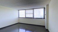 This single-floor space presents a straightforward and functional layout with a clean, minimalist aesthetic. The room is defined by a row of large windows fitted with horizontal metal blinds, allowing ample natural light to filter in while offering views of the surrounding urban environment, including neighboring multi-story buildings. The windows span a generous portion of the exterior wall, contributing to an open and airy atmosphere despite the compact proportions of the room.

The flooring consists of polished dark tiles that reflect incoming light, adding a sleek, professional quality to the space. The walls are finished in a neutral off-white tone, providing a simple and unobtrusive backdrop that complements the overall utilitarian character of the interior. The ceiling features a dark textured finish, which contrasts with the lighter walls and introduces a subtle visual depth to the room.

Black baseboard trim runs along the perimeter, creating a defined boundary between the walls and the highly reflective floor. The space is currently unfurnished, making it highly versatile and adaptable for a range of uses. Overall, the interior conveys a functional, professional ambiance suited to commercial or office-style occupancy.
