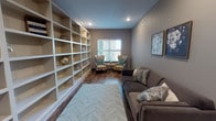 This single-floor library is a well-appointed space designed to balance functionality with comfort. The room's most striking feature is the expansive floor-to-ceiling built-in shelving unit that spans the full length of one wall, offering generous storage and display capacity for books, décor, and collectibles. The shelving is finished in crisp white, providing a clean contrast against the warm, neutral gray walls that define the room's calm and focused atmosphere.

Durable hardwood-style flooring in rich, warm tones runs throughout the space, anchoring the room with a classic and inviting aesthetic. A herringbone-patterned area rug adds texture and visual interest beneath the central seating area. Comfortable lounge seating, including a plush gray sofa and a pair of floral wingback chairs, creates an ideal environment for relaxed reading or informal conversation. A compact side table nestled between the chairs provides a practical surface near the window.

Natural light filters in through a window fitted with blinds, offering a view to the outdoors. Recessed ceiling lighting supplements natural light throughout the day. Toward the entrance, a small study table with chairs provides a dedicated workspace. Glass-paned French doors offer a transparent boundary between the library and the adjacent corridor, maintaining an open and connected feel while defining the space clearly.