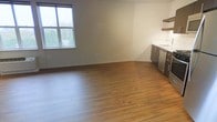 This single-floor space offers a clean, modern aesthetic defined by warm hardwood-style flooring that extends throughout the open layout, creating a sense of continuity and spaciousness. The living area is bathed in natural light, courtesy of a large multi-pane window that frames a lush, green outdoor view — a standout feature that adds life and brightness to the interior. The neutral, light-toned walls complement the warm flooring tones, creating a welcoming and versatile environment.

Transitioning seamlessly from the living area, the kitchen is efficiently designed with dark, contemporary cabinetry paired with a crisp white countertop and a stainless steel sink. An open floating shelf above provides practical storage while maintaining the space's airy feel. A stainless steel dishwasher is integrated into the cabinetry, adding a modern, functional touch. The open-plan arrangement between the living and kitchen zones makes the space feel expansive and well-connected, ideal for both relaxed everyday living and entertaining. Overall, the space strikes a balance between modern simplicity and functional design.
