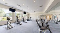 Located at 31220 Samantha Ln in Temecula, CA, this well-appointed fitness center offers a single, generously sized exercise room designed to support a comprehensive workout experience. The space is bathed in natural light, thanks to a large bank of floor-to-ceiling windows along one wall that frame views of surrounding greenery, creating an inviting and energizing atmosphere.

The exercise room is thoughtfully equipped with a wide range of cardio machines, including treadmills, elliptical trainers, and a stationary bike, all positioned to take advantage of the natural light and outdoor views. A dedicated strength-training area features multiple cable and weight machines, along with a dumbbell rack, catering to a full range of resistance workouts.

Full-length mirrors line one wall, visually expanding the space while allowing users to monitor their form during exercise. Overhead, recessed lighting supplements the abundant natural light, ensuring consistent illumination throughout the room. Wall-mounted flat-screen televisions are strategically placed for viewing during cardio sessions. The floor is finished with durable, patterned carpet tiles that provide comfort and traction underfoot. The neutral color palette of the walls and ceiling contributes to a clean, modern aesthetic that keeps the focus on fitness.