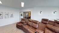 This single-floor communal lounge space combines comfort and modern industrial design in a thoughtfully arranged layout. The media area serves as the centerpiece, furnished with plush brown leather recliner seating arranged in a theater-style configuration, ideal for group viewing and relaxation. A wall-mounted flat-screen TV is positioned across from the seating, accompanied by a rustic wood media console with decorative accents.

The space is characterized by light hardwood-style flooring, crisp white walls, and exposed black ductwork along the ceiling, lending an urban, industrial aesthetic. Recessed and flush-mounted ceiling lights, complemented by wall-mounted lantern-style sconces, provide warm, layered illumination throughout.

A bistro-style nook features a round wood-top table with leather bar stools positioned directly in front of a wide bank of windows, offering views of the surrounding outdoor landscape — an inviting spot for casual conversation or individual use.

Large dark-framed glass partition walls and a windowed interior door create a visual connection to an adjacent room, enhancing the open, airy feel of the space. A bold mixed-media artwork piece adds a creative focal point near the window nook, contributing to the space's contemporary character.