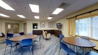 This community and function room offers a well-appointed, single-level space designed to accommodate a variety of gatherings and events. The room features a drop ceiling with recessed panel lighting that provides even, bright illumination throughout. The flooring consists of a patterned carpet in neutral blue and tan tones, adding a polished yet welcoming aesthetic.

Seating is arranged flexibly, with multiple round tables accompanied by blue stacking chairs, as well as a pair of cushioned armchairs that offer a more relaxed seating option. Along the perimeter, dark wood cabinetry with lower storage units provides ample surface space and storage, suitable for serving refreshments or organizing materials.

A wall-mounted flat-screen television is positioned for easy viewing from most areas of the room, making the space practical for presentations or casual viewing. Framed artwork on the walls contributes a refined touch to the overall decor. A window dressed with warm-toned drapes introduces natural light while offering privacy when needed. The room's neutral color palette and functional layout make it adaptable for community meetings, social events, and organized functions alike.