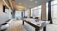 Located at 215 N Pine Street in the heart of uptown Charlotte, NC, this contemporary amenity lounge is designed for both recreation and relaxed socializing. The space is anchored by floor-to-ceiling windows that flood the interior with natural light and offer impressive views of the Charlotte skyline, creating a dynamic urban backdrop throughout the day. A sophisticated geometric-patterned carpet runs the length of the room, tying together the modern aesthetic with a subtle city-map motif. The lounge is well-equipped for entertainment, featuring a full-size billiards table as its centerpiece, complemented by a foosball table and a ping-pong table. An eye-catching sputnik chandelier adds a bold design statement overhead, while recessed lighting provides additional illumination. Plush armchairs with chrome frames are positioned near the windows, offering comfortable seating with panoramic city views. A dedicated lounge nook features a wall-mounted flat-screen TV, acoustic wall panels, and sleek metallic sphere seating alongside bistro-style bar tables. Bold abstract artwork accents the walls throughout, adding an artistic flair to the space. The overall ambiance is polished, vibrant, and distinctly urban.