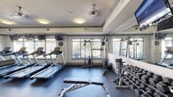 Located at 8500 Franciscan Woods Dr in Columbus, Georgia, this fitness center offers a comprehensive workout environment within a thoughtfully designed single-floor space. The room features a tray ceiling with crown molding trim, ceiling fans, and flush-mount light fixtures that create a bright, comfortable atmosphere. Large multi-pane windows line the walls on multiple sides, flooding the interior with natural light and offering views of the lush, tree-lined outdoor surroundings. The durable rubber flooring throughout provides both safety and functionality for a variety of exercises. The space is well-stocked with cardio equipment, including multiple treadmills and an elliptical machine, as well as a stationary bike. Strength training needs are addressed with a cable weight machine, a weight bench, a squat rack, and a full dumbbell rack with an assorted range of free weights. Wall-mounted flat-screen televisions are positioned above the cardio machines for entertainment during workouts. A large wall mirror expands the visual depth of the space and assists with exercise form. A decorative column adds an architectural accent near the center of the room. The overall ambiance is clean, functional, and welcoming, making it a practical fitness facility.