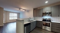 Located at 1001 National Ave in San Bruno, California, this single-floor residence offers a well-designed layout with modern finishes throughout. The open-concept living and kitchen area serves as the heart of the home, featuring rich dark hardwood-style flooring, neutral gray walls, and track lighting that adds a contemporary touch. Natural light filters in through windows, enhancing the bright and airy atmosphere.

The kitchen is thoughtfully appointed with gray flat-front cabinetry, white quartz countertops, and a classic white subway tile backsplash. Stainless steel appliances — including a refrigerator, range, microwave, and dishwasher — are integrated seamlessly into the design. A peninsula counter with a built-in sink provides additional prep space and serves as a natural divider between the kitchen and living area.

The bedrooms are finished with soft carpeting and include a wall-mounted mini-split HVAC unit for individual climate control, along with windows that bring in natural light and views of the surrounding greenery. Closet access is conveniently located within the bedrooms.

The bathroom features a vanity with gray cabinetry and white countertop, a tub-shower combination with white tile surround, and hardwood-style flooring, maintaining the cohesive aesthetic found throughout the residence.
