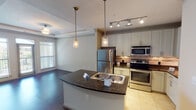 Located at 5201 Collin-McKinney Pkwy in McKinney, TX, this single-floor residence offers a thoughtfully designed layout with a seamless blend of style and functionality. The open-concept kitchen serves as a central highlight, featuring dark granite countertops, a curved island with a stainless steel sink, white cabinetry with crown molding, a stone tile backsplash, and a full suite of stainless steel appliances including a refrigerator, electric range, and microwave. Track lighting and elegant pendant lights illuminate the kitchen, which flows naturally into the living and dining areas. The living area boasts rich dark hardwood flooring and a tray ceiling with detailed crown molding, adding architectural character. A ceiling fan and large windows with blinds provide natural light and ventilation. The bedrooms offer plush carpeting, ceiling fans, and windows that bring in ample daylight. The primary bedroom is accessed through stately double doors. The bathroom is finished with granite-style countertops, dual sinks, white cabinetry, and a tiled shower/tub combination. Light tile flooring in the kitchen transitions smoothly to dark hardwood in the main living spaces, creating distinct yet cohesive zones throughout the residence.