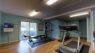 This fitness center is a dedicated single-floor exercise space designed to support a full range of workout routines. The room features warm hardwood flooring complemented by protective rubber mat sections strategically placed beneath the equipment to ensure safety and stability during use. The space is outfitted with a multi-function cable weight machine, a Life Fitness treadmill, and a rowing or bench press station, providing options for both cardiovascular and strength training workouts. Soft teal-green walls lend a calm, energizing atmosphere to the room, while bright fluorescent lighting ensures excellent visibility throughout. A large sliding glass door along one wall floods the space with natural light and offers a view of a landscaped outdoor area, creating an open and inviting feel despite the compact footprint. Instructional fitness posters are mounted on the walls, adding a functional and informative touch to the environment. The layout is thoughtfully arranged to allow comfortable movement between equipment. Overall, this fitness center offers a practical and well-maintained environment suitable for a variety of fitness needs.