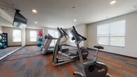 Located at 4604 Hammersley Road in Madison, Wisconsin, this fitness center is a single-floor facility designed to support a well-rounded exercise routine. The space is outfitted with a selection of Precor cardio equipment, including treadmills, elliptical trainers, and a stationary bike, arranged to allow comfortable movement between machines. A dedicated strength training area features a multi-tiered dumbbell rack and a weight bench, providing options for resistance-based workouts. A rack of exercise balls in varying sizes adds versatility for functional fitness and stretching routines. A large wall-mounted mirror spans one side of the room, visually expanding the space and allowing users to monitor their form during exercise. The flooring consists of patterned carpet tiles in warm orange and gray tones, adding both comfort underfoot and visual character to the room. Multiple windows along the walls invite abundant natural light, creating a bright and energizing atmosphere. Recessed ceiling lighting supplements the daylight, ensuring consistent illumination throughout all hours. The neutral-toned walls and clean layout contribute to an organized, focused environment suited for daily fitness use.
