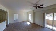 This single-floor space presents an open and functional layout centered around a generously sized main living area finished with wall-to-wall carpeting in a neutral tone. The room is illuminated by a combination of natural light streaming through a centrally positioned window and two overhead light fixtures — a classic multi-blade ceiling fan and a simple pendant light that hangs at a comfortable height, adding a subtle focal point to the space. The neutral, light-toned walls throughout create a clean and versatile backdrop suitable for a variety of interior styles. A doorway visible from the main area leads to an adjacent room, also carpeted, suggesting a connected layout that supports easy flow between spaces. On the opposite side, a closet or storage area can be identified, contributing to the practicality of the floor plan. The overall ambiance is understated and move-in ready, with a calm, neutral palette that allows for flexible personalization. The space benefits from a straightforward single-level design, making it accessible and easy to navigate throughout.