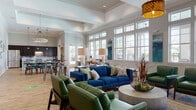 Located at 100 Shellbark Way in Savannah, Georgia, this single-story community clubhouse offers a well-designed gathering space that blends comfort and functionality. The open-plan layout is anchored by a spacious lounge area featuring coffered ceilings, recessed lighting, and rich hardwood-style flooring throughout. Large, multi-pane windows line the walls, flooding the interior with natural light and offering views of the surrounding community and pool area.

The seating areas are thoughtfully arranged with vibrant blue velvet sofas, green leather accent chairs, and mid-century modern furnishings that create a welcoming, resort-style atmosphere. A built-in banquette with bold blue-and-white striped wall paneling and wood wainscoting provides an intimate dining nook, complemented by pendant lighting and patterned upholstery.

The fully equipped kitchen features dark cabinetry extending to the ceiling, a granite-topped island with bar seating, stainless steel appliances including a full-size refrigerator, a mosaic tile backsplash, and pendant lights overhead. High-top bar tables positioned along the window walls offer casual seating with scenic outdoor views. Curated coastal artwork and layered lighting complete the polished, inviting ambiance throughout the space.