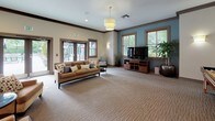 This single-floor community clubhouse is thoughtfully designed to offer residents a comfortable and multifunctional gathering space. The open layout is anchored by a generously sized lounge area with plush seating, a wall-mounted television, and warm pendant lighting overhead. Neutral carpet flooring, crown molding, and a soothing palette of soft blue and beige tones create a welcoming ambiance throughout.

The fully equipped kitchen is a standout feature, outfitted with rich wood cabinetry, granite countertops, a mosaic tile backsplash, stainless steel appliances, and recessed lighting. A built-in breakfast bar with bar-stool seating connects the kitchen to the main gathering area, making it ideal for social events and community functions.

A cozy dining nook, accented by soft blue walls and a window with natural light, provides an intimate setting for smaller gatherings. Adjacent to the main lounge, a dedicated game room houses a full-length shuffleboard table, adding a recreational dimension to the space.

Large glass doors and windows along one wall flood the interior with natural light and open directly to a furnished outdoor patio, offering seamless indoor-outdoor connectivity. The overall design balances practicality with comfort, making this clubhouse a versatile and inviting hub for community life.