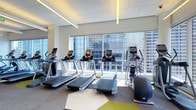 This fitness center offers a well-appointed workout environment designed with both functionality and aesthetics in mind. The space is characterized by expansive floor-to-ceiling windows that span the length of the cardio equipment area, providing sweeping views of the surrounding urban skyline and flooding the interior with natural light. The bright, open atmosphere is further enhanced by a clean white drop ceiling accented with linear LED lighting strips that add a modern, polished touch.

The cardio zone is prominently arranged along the window wall and features a row of modern treadmills equipped with individual digital displays, alongside an elliptical trainer, allowing users to exercise while enjoying the city views. The flooring incorporates a geometric-patterned carpet with bold yellow-green and grey tones, adding a dynamic visual element to the space.

The overall design balances a contemporary aesthetic with practical layout considerations, ensuring equipment is accessible and well-spaced. The combination of abundant natural light, modern finishes, and high-rise views creates an energizing environment well-suited for an effective workout experience.