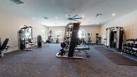 This fitness center is a comprehensive, single-floor facility designed to support a full range of workout routines. The space is finished with durable patterned carpet flooring and neutral-toned walls, creating a clean and functional environment throughout. Recessed lighting is complemented by large windows along multiple walls, allowing natural light to fill the room and providing views of the surrounding outdoor greenery.

The cardio zone features a row of treadmills, each equipped with individual screens, alongside elliptical trainers, upright bikes, and stair climbers — offering a well-rounded selection for aerobic exercise. A wall-mounted flat-screen television is positioned to keep users engaged during cardio sessions.

The strength training area is equally well-appointed, housing a cable crossover machine, weight benches, a power rack with barbell and plates, and a full dumbbell rack with a wide range of weights. Dedicated strength machines, including leg press and leg extension/curl units, are situated in a quieter alcove near a glass-paneled door that overlooks an adjacent outdoor area.

A large ceiling fan provides additional air circulation, and rubber floor mats are placed in high-use lifting zones for safety. The overall layout is spacious and thoughtfully organized, ensuring smooth traffic flow between exercise stations throughout the facility.