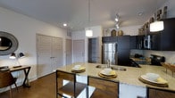 This single-story residence at Breakwater showcases a thoughtfully designed open-concept kitchen that serves as the focal point of the living space. The kitchen is anchored by a spacious island featuring granite countertops with a double undermount sink, providing ample workspace and a natural gathering spot. Industrial-style bar stools with wood shelf detailing complement the island's clean lines, adding a touch of rustic contrast to the contemporary design.

Dark espresso cabinetry lines the kitchen walls, offering generous storage while creating a bold visual statement against the neutral white walls and light gray ceiling. A stainless steel refrigerator anchors the appliance suite, which is further accentuated by track lighting and a pendant light above the island, ensuring well-distributed illumination throughout the space. An open shelving unit provides convenient additional storage and display space.

The warm hardwood flooring runs throughout, adding richness and continuity. Multiple doorways visible from the kitchen suggest a well-connected floor plan with easy access to adjoining rooms. Recessed lighting complements the overall bright, clean aesthetic, making this a highly functional and visually appealing living space.