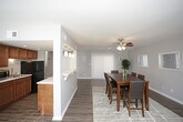 This single-floor space is designed with an open-concept layout that promotes a seamless flow between the kitchen, dining, and living areas. Warm hardwood-style flooring runs consistently throughout, tying the rooms together with a cohesive and inviting aesthetic. The walls are finished in a soft, neutral gray tone that enhances the brightness of the interior.

The kitchen is thoughtfully arranged in a galley-style configuration, featuring rich warm-toned wood cabinetry, light countertops, a stainless steel sink, and black appliances including a full-size refrigerator. Recessed lighting provides focused illumination, and a pass-through opening connects the kitchen directly to the adjacent living and dining spaces.

The dining area is generously sized and anchored by a ceiling fan with integrated lighting. A large sliding glass door dressed with vertical blinds allows natural light to fill the room, creating an airy atmosphere. Decorative framed mirrors along the wall add depth to the space.

The living area is a comfortable, well-proportioned room with a large window fitted with horizontal blinds that offer both natural light and privacy. A wall-mounted pass-through window maintains a visual connection to the kitchen. Together, the spaces form a functional and well-balanced single-floor layout.