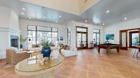 This single-story community clubhouse is thoughtfully designed to offer a range of amenities within a cohesive, light-filled environment. Throughout the space, warm terracotta tile flooring, crisp white walls, and black-framed windows establish a consistent and inviting aesthetic.

The main lounge area is anchored by soaring ceilings and generous wall-to-wall windows fitted with French doors that open to an outdoor patio. Comfortable white upholstered seating is arranged around a glass-topped coffee table, creating an open and welcoming gathering space. Coastal-inspired artwork and decorative accents contribute to the relaxed ambiance.

Adjacent to the lounge, the game room features a full-size pool table and tall bistro-style tables with bar seating, with direct views and access to the outdoor area through steel-framed glass doors.

The kitchenette is well-appointed with rich dark wood cabinetry, granite countertops, a central island with bar seating, stainless steel appliances, and a striking arched window that frames outdoor views and fills the room with natural light.

The fitness center is divided into two well-equipped areas—one dedicated to cardio machines including treadmills and stationary bikes, and another to strength training with cable machines, free weights, and floor-to-ceiling mirrors. Dual water fountains are conveniently located nearby.