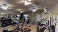 This single-story fitness center offers a practical and functional space designed to support a range of exercise routines. The room features hardwood-style flooring throughout, providing a clean and durable surface suited for gym use. Multiple ceiling fans are installed overhead, ensuring adequate air circulation and comfort during workouts.

The space is outfitted with a solid selection of exercise equipment, including a multi-station strength training tower, a recumbent stationary bike, an elliptical machine, a treadmill, and various weight benches and resistance machines. This variety accommodates both cardiovascular and strength-focused training.

Natural light enters through windows fitted with blinds along one wall, supplemented by overhead fluorescent lighting to keep the space well-lit at all times. A large wall mirror on one side visually expands the room and aids users in monitoring their form during exercise. Motivational fitness posters are displayed on the walls, contributing to the gym atmosphere. A clearly marked exit door with illuminated signage ensures safety and easy access. The overall ambiance is practical, clean, and focused on utility.