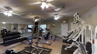 This single-story fitness center offers a practical and functional space designed to support a range of exercise routines. The room features hardwood-style flooring throughout, providing a clean and durable surface suited for gym use. Multiple ceiling fans are installed overhead, ensuring adequate air circulation and comfort during workouts.

The space is outfitted with a solid selection of exercise equipment, including a multi-station strength training tower, a recumbent stationary bike, an elliptical machine, a treadmill, and various weight benches and resistance machines. This variety accommodates both cardiovascular and strength-focused training.

Natural light enters through windows fitted with blinds along one wall, supplemented by overhead fluorescent lighting to keep the space well-lit at all times. A large wall mirror on one side visually expands the room and aids users in monitoring their form during exercise. Motivational fitness posters are displayed on the walls, contributing to the gym atmosphere. A clearly marked exit door with illuminated signage ensures safety and easy access. The overall ambiance is practical, clean, and focused on utility.