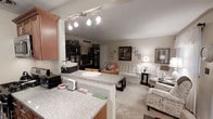 This single-floor residence showcases an open-concept design that seamlessly integrates the kitchen and living areas into one cohesive, functional space. The kitchen features granite countertops with a speckled grey-and-white finish, wood-toned cabinetry, and a peninsula that serves as both a workspace and an informal divider between the kitchen and the living area. A modern track lighting fixture overhead provides focused illumination across the kitchen surfaces.

The transition into the living area is natural and unobstructed, creating a sense of openness and ease of movement. Soft, neutral-toned carpeting covers the living room floor, contributing to a warm and comfortable ambiance. The living space is well-appointed, featuring seating with classic upholstery, decorative side tables, and framed artwork displayed along the walls, lending the room a curated, homey character.

Neutral wall tones are carried throughout both spaces, creating visual consistency and a calm atmosphere. The combination of track lighting in the kitchen and warm lamp lighting in the living room offers layered illumination suited to both task-oriented and relaxed settings. Overall, the space presents a practical yet inviting layout designed for comfortable everyday living.