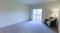 This single-floor residential space offers a clean, functional layout designed for comfortable everyday living. The main living area is generously sized, featuring soft wall-to-wall carpeting in a neutral blue-gray tone that adds warmth and comfort underfoot. Smooth, light-colored walls provide a calm and airy backdrop throughout the space, complemented by a ceiling with a subtle height that contributes to an open, uncluttered feel. A standout feature of the living area is the large sliding glass door that fills the room with abundant natural light, creating a bright and welcoming atmosphere. The door opens directly onto a private balcony enclosed by a metal railing, offering views of the surrounding exterior environment. The transition between the interior and the outdoor space is seamless, making the balcony a natural extension of the living area. Recessed ceiling vents suggest a central HVAC system for year-round climate comfort. The overall ambiance of the space is understated and versatile, with neutral finishes that provide an excellent canvas for a variety of interior styles and furnishing arrangements.