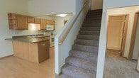 This two-story residence is designed with functionality and comfort in mind. The main level showcases a well-equipped kitchen featuring warm honey-toned wood cabinetry, a double sink, electric range, and built-in microwave. A peninsula with a decorative panel provides additional counter space and a casual dining or prep area, complemented by dark stone-look countertops that add a grounding contrast to the light wood tones. The flooring on the main level is light-toned hardwood or laminate, creating a bright and clean aesthetic throughout.

A carpeted staircase with a wood handrail connects the main level to the upper floor. The stairs are finished in a neutral-toned, textured carpet that continues to provide comfort underfoot. The stairwell is open and well-lit, contributing to an airy feel in the transitional space between floors.

The overall interior palette is neutral and understated, with light gray walls and warm wood accents throughout. The layout makes efficient use of space, with clearly defined areas for cooking, living, and circulation. The two-floor configuration offers a natural separation between living and private areas, making this a practical and well-organized home.