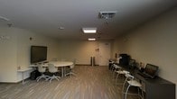 This single-floor space is thoughtfully designed to support both collaborative work and individual productivity. The main area features a central oval conference table surrounded by ergonomic rolling chairs, making it well-suited for team meetings and group discussions. A large flat-screen display mounted on the wall serves as a focal point for presentations and visual collaboration.

The space is finished with warm-toned wood-look flooring that adds a professional yet approachable feel throughout. Overhead, recessed and flush-panel lighting fixtures provide consistent, even illumination across the room. A ceiling-mounted projector further enhances the room's presentation capabilities.

Along one side of the room, a row of high bar-style workstations with stools offers an alternative seating arrangement for breakout work or informal collaboration. This area also includes computer equipment and what appears to be a printer or multifunction device, supporting everyday office tasks.

The neutral-toned walls and clean, uncluttered layout contribute to a focused and functional ambiance. A single interior door provides access to adjacent areas, while HVAC vents in the ceiling ensure climate control throughout. Overall, the space strikes a balance between meeting room functionality and practical workspace utility.