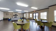 This single-level community room is thoughtfully designed to accommodate a variety of gatherings and social activities. The space is divided into distinct functional zones, each contributing to an inviting and versatile environment. Dark hardwood-style flooring runs throughout, providing a cohesive foundation that complements the warm, neutral wall tones accented by dark crown molding.

The lounge area serves as a central gathering point, anchored by a tiled fireplace with a dark mantel and flanked by built-in bench seating with colorful accent pillows. Decorative wall art and an area rug add personality, while mid-century-style chairs and ottomans create a relaxed, comfortable atmosphere.

The dining area features multiple round tables surrounded by chartreuse upholstered chairs, offering ample seating for group meals or events. Large windows and glass doors allow natural light to fill the space and provide views to the exterior.

The kitchen area is well-equipped with dark cabinetry, light countertops, a full-size refrigerator, microwave, and sink. A peninsula counter provides additional prep and serving space, making it practical for catered events or community meals. Overhead fluorescent lighting ensures consistent, functional illumination throughout the entire room.