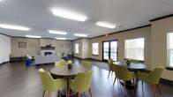 This single-level community room is thoughtfully designed to accommodate a variety of gatherings and social activities. The space is divided into distinct functional zones, each contributing to an inviting and versatile environment. Dark hardwood-style flooring runs throughout, providing a cohesive foundation that complements the warm, neutral wall tones accented by dark crown molding.

The lounge area serves as a central gathering point, anchored by a tiled fireplace with a dark mantel and flanked by built-in bench seating with colorful accent pillows. Decorative wall art and an area rug add personality, while mid-century-style chairs and ottomans create a relaxed, comfortable atmosphere.

The dining area features multiple round tables surrounded by chartreuse upholstered chairs, offering ample seating for group meals or events. Large windows and glass doors allow natural light to fill the space and provide views to the exterior.

The kitchen area is well-equipped with dark cabinetry, light countertops, a full-size refrigerator, microwave, and sink. A peninsula counter provides additional prep and serving space, making it practical for catered events or community meals. Overhead fluorescent lighting ensures consistent, functional illumination throughout the entire room.