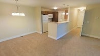 This single-floor residence features an open-concept design that seamlessly connects the kitchen and living area into one cohesive space. The living area is finished with wall-to-wall neutral carpeting, contributing to a warm and understated ambiance throughout. Soft, cream-colored walls enhance the brightness of the space.

The kitchen is equipped with white appliances, including a refrigerator, microwave, and stovetop, complemented by rich dark brown cabinetry that provides ample storage. A peninsula-style breakfast bar serves as a natural dividing element between the kitchen and the living area, adding both functional counter space and a casual dining option. Two pendant lights hang above the bar, providing focused task lighting and a subtle decorative touch.

Recessed and flush-mounted ceiling fixtures offer consistent ambient lighting throughout the space. The open layout allows for flexible furniture arrangement in the living area, making the space feel adaptable and functional. Overall, the unit presents a clean, move-in ready environment with a practical and straightforward design aesthetic.