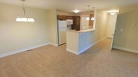 This single-floor residence features an open-concept design that seamlessly connects the kitchen and living area into one cohesive space. The living area is finished with wall-to-wall neutral carpeting, contributing to a warm and understated ambiance throughout. Soft, cream-colored walls enhance the brightness of the space.

The kitchen is equipped with white appliances, including a refrigerator, microwave, and stovetop, complemented by rich dark brown cabinetry that provides ample storage. A peninsula-style breakfast bar serves as a natural dividing element between the kitchen and the living area, adding both functional counter space and a casual dining option. Two pendant lights hang above the bar, providing focused task lighting and a subtle decorative touch.

Recessed and flush-mounted ceiling fixtures offer consistent ambient lighting throughout the space. The open layout allows for flexible furniture arrangement in the living area, making the space feel adaptable and functional. Overall, the unit presents a clean, move-in ready environment with a practical and straightforward design aesthetic.
