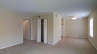 This single-floor residence offers a straightforward and functional layout designed for comfortable everyday living. The main living area is finished with wall-to-wall carpeting and neutral, warm-toned walls that create a clean and understated ambiance throughout. A ceiling fan is installed in the living space, providing both light and airflow. Crown molding details add a subtle architectural touch to the rooms.

A central structural wall divides the living area from the kitchen zone, which is visible in the background and features standard appliances, including a refrigerator. The transition between spaces feels open and accessible, lending the floor plan a connected, airy quality.

A dedicated storage area includes a set of bifold closet doors that open to reveal a built-in closet with additional shelving or rod space, offering practical organization solutions. A secondary door nearby leads to an adjacent room or hallway, further suggesting a well-connected interior layout.

HVAC vents are positioned thoughtfully in the ceiling, ensuring climate control throughout the unit. The overall design is simple and move-in ready, with consistent finishes across all visible spaces that emphasize functionality and ease of living.