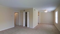This single-floor residence offers a straightforward and functional layout designed for comfortable everyday living. The main living area is finished with wall-to-wall carpeting and neutral, warm-toned walls that create a clean and understated ambiance throughout. A ceiling fan is installed in the living space, providing both light and airflow. Crown molding details add a subtle architectural touch to the rooms.

A central structural wall divides the living area from the kitchen zone, which is visible in the background and features standard appliances, including a refrigerator. The transition between spaces feels open and accessible, lending the floor plan a connected, airy quality.

A dedicated storage area includes a set of bifold closet doors that open to reveal a built-in closet with additional shelving or rod space, offering practical organization solutions. A secondary door nearby leads to an adjacent room or hallway, further suggesting a well-connected interior layout.

HVAC vents are positioned thoughtfully in the ceiling, ensuring climate control throughout the unit. The overall design is simple and move-in ready, with consistent finishes across all visible spaces that emphasize functionality and ease of living.