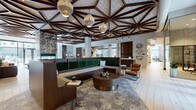 This expansive amenity lobby is defined by its remarkable geometric ceiling, where dark wood beams form an intricate star-burst lattice pattern across the soaring overhead expanse, punctuated by stylish pendant light fixtures. Light wood-plank flooring runs throughout, lending warmth and continuity across the open-plan layout.

The central lounge area anchors the space with a large L-shaped green velvet banquette, paired with rounded wood and brass coffee tables and cognac leather accent chairs arranged atop layered patterned rugs. Flowing into an adjacent clubroom, a well-appointed bar counter features marble surfaces, woven pendant lights, blue velvet seating, and botanical wallcovering, creating an inviting social hub. Wall-mounted screens and varied seating arrangements offer flexible gathering options nearby.

A glass-enclosed private office provides a functional workspace visible from the main corridor, while a dedicated mail room features floor-to-ceiling dark wood parcel lockers and mailboxes with clean marble countertops. Decorative wall plates and carved wood screens add artistic detail throughout. Floor-to-ceiling windows and glass doors at one end flood the space with natural light and provide direct access to an outdoor patio courtyard, seamlessly connecting interior and exterior living.