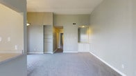 This single-floor space presents a spacious, open living area finished in soft sage-green and warm neutral tones throughout. The main living area is carpeted in light grey, contributing to a calm, understated atmosphere. Notably tall walls lend an open, airy feel to the room, enhancing the sense of volume beyond its footprint. A built-in shelving unit is recessed into one wall, providing organized display or storage space, while a mirrored niche with a lit interior and white cabinetry below serves as a functional and visually appealing accent on the opposite wall. A hallway visible in the background suggests additional rooms beyond, with a doorway leading further into the unit. Consistent white baseboards frame the perimeter of the room, complementing the clean, well-maintained aesthetic. A ceiling vent is visible, indicating a central HVAC system. The overall design is simple and practical, with built-in features that maximize utility without sacrificing the open character of the main living space.